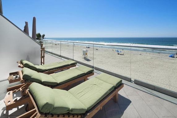 Beachfront view from a terrace with lounge chairs and glass railing.