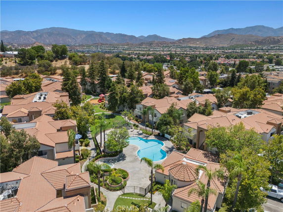 Panoramic view of residential area with multiple houses and a central swimming pool, surrounded by trees and distant mountains.