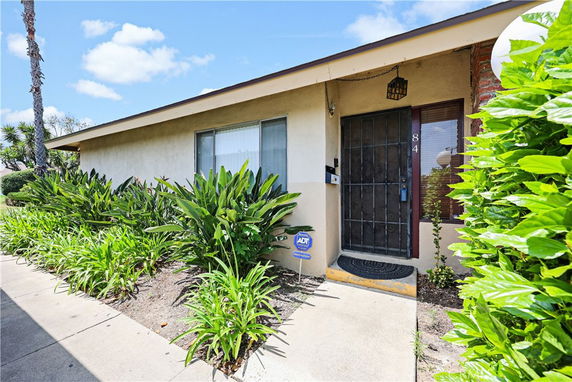 Front view of a single-story house with a metal security door and surrounding greenery.