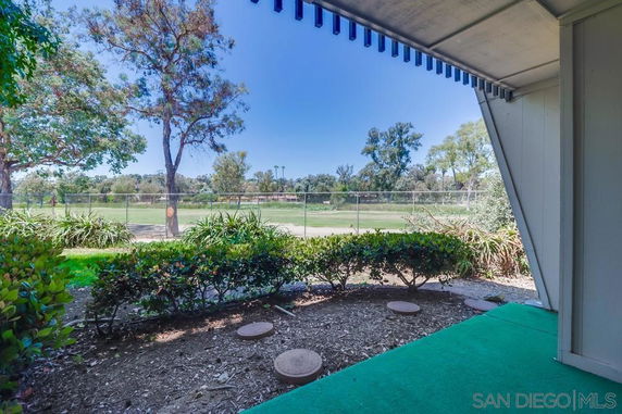 View from under an awning showing a garden area with shrubs and trees, overlooking a grassy field with a fence.