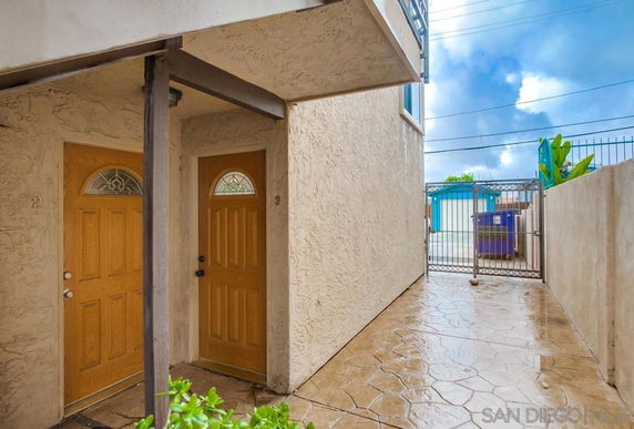 Ground-level view of two wooden entrance doors in a stucco building with a narrow passage leading to an outdoor gate.