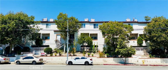 Front view of a multi-story apartment building with balconies and a line of trees and cars parked in front.