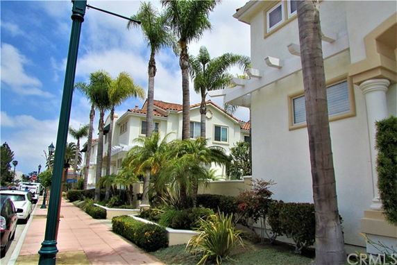 Street view of a row of multi-story houses with balconies and palm trees lining the sidewalk.