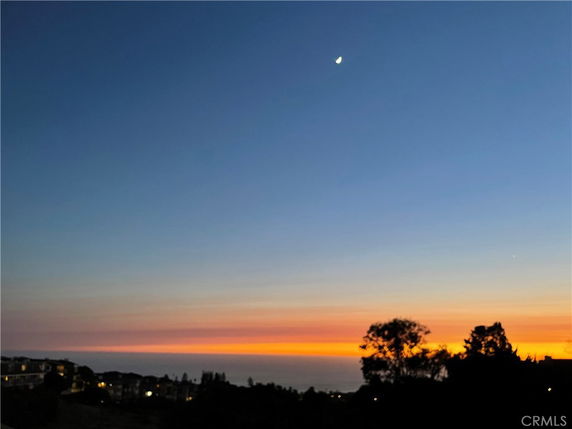 Panoramic view of the horizon at dusk with the moon visible in the sky and silhouettes of distant buildings and trees.