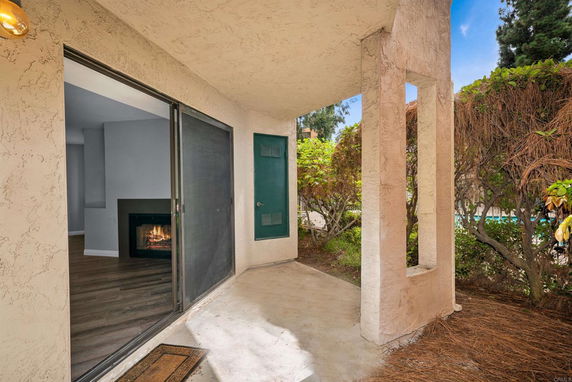 Covered patio outside a house with sliding glass door leading to a living room with a fireplace. Bushes and greenery are visible.
