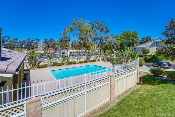 Outdoor swimming pool surrounded by a fence with nearby buildings and trees.