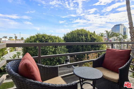 View from a balcony with two chairs and a small table overlooking trees and a cityscape.