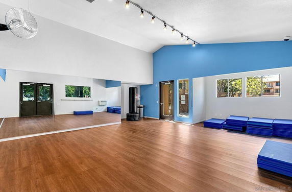 Interior view of a recreational room with wooden flooring, blue accented walls, exercise mats, and mirrors on the wall.