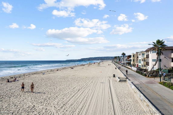 Beachfront view with sandy shore, ocean waves, and adjacent residential buildings.