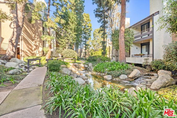 Outdoor view with a small stream, bridge, and trees surrounding a residential building.