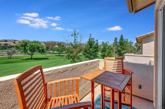 A small balcony with wooden chairs and table overlooking a green golf course and trees.