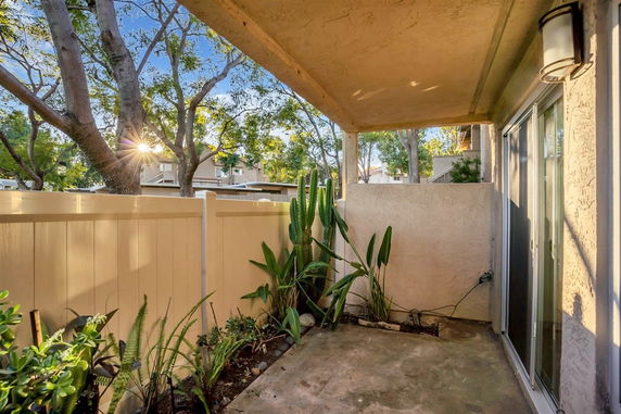 Covered outdoor patio area with plants and a fence, adjacent to a building.