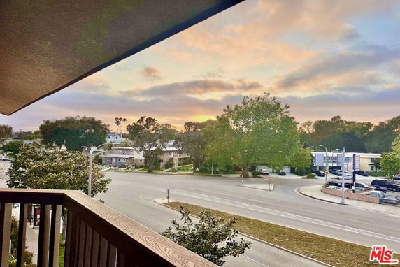 View from a balcony overlooking a street and surrounding buildings with trees and a sunset sky.