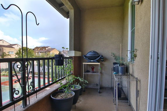 A balcony area with potted plants and a small grill, overlooking nearby buildings and a sports court.