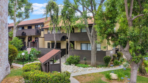 Front view of a two-story residential building with balconies and a brown exterior, surrounded by trees and landscaped area.