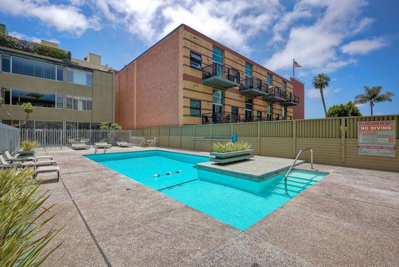 Outdoor swimming pool area with loungers beside a multi-story building.
