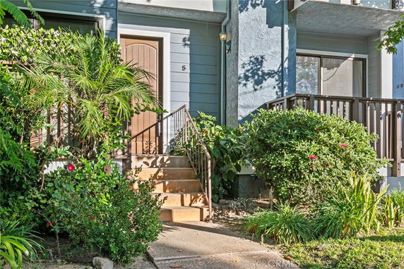 Front view of a townhouse with a wooden door and steps leading to the entrance, surrounded by plants.