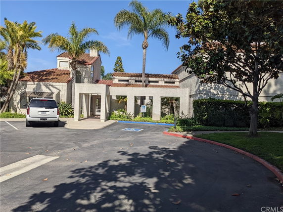Front view of a building with a tile roof and entryway, featuring palm trees and a parking area.