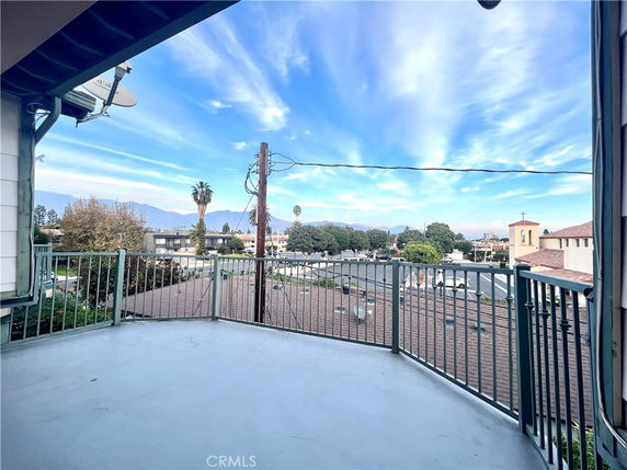 Panoramic view from a balcony overlooking rooftops and trees, with distant mountains visible under a blue sky.