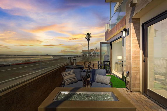 View of a modern building's outdoor seating area by the seaside, with glass doors and a sunset backdrop.