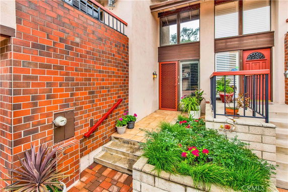 Front view of a house with a red brick facade and entryway steps, featuring potted plants and a red-painted door.