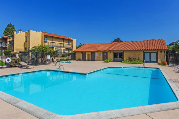 Outdoor swimming pool area with a tan building featuring a red tile roof and fencing around the pool.