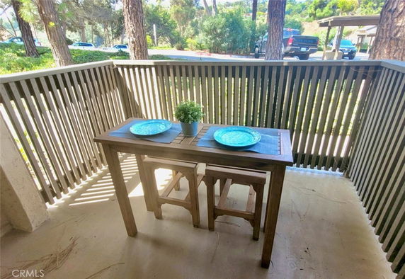 Small outdoor balcony with a wooden table and two stools, overlooking trees and parked vehicles.