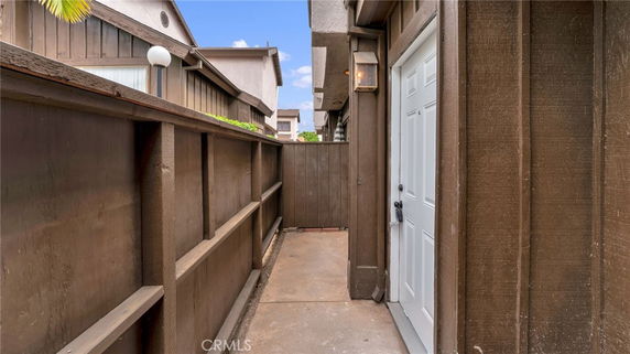 View of a narrow walkway next to a wooden fence leading to a building entrance with a white door.