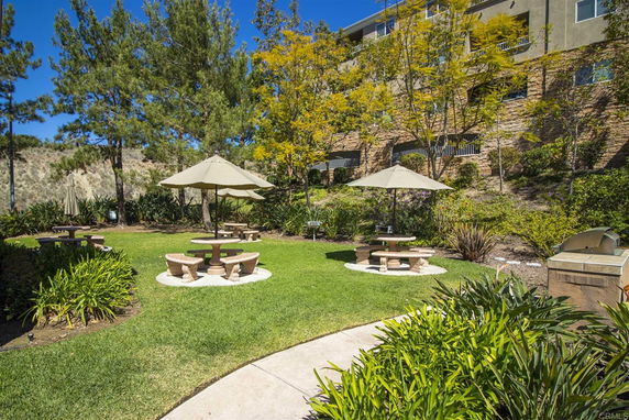 Exterior view of a garden area with picnic tables and umbrellas, surrounded by trees and a building in the background.