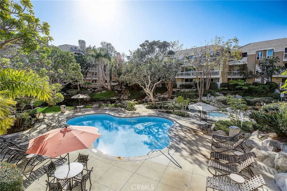 Outdoor area with a swimming pool surrounded by lounge chairs, tables with umbrellas, and landscaped greenery.