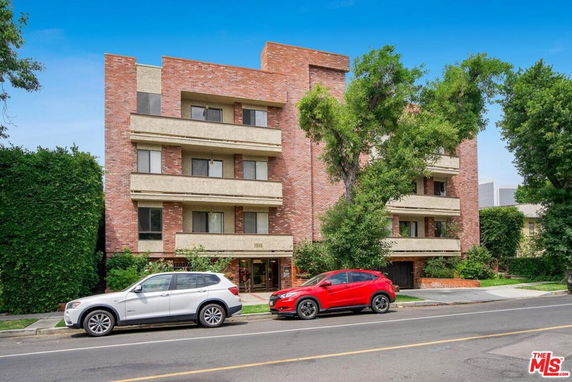 Front view of a multi-story brick apartment building with balconies and a parking area in front.