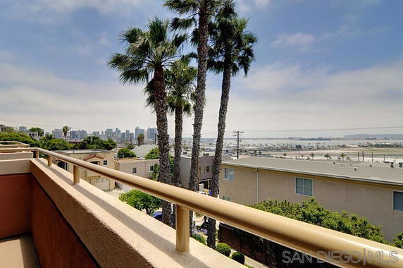 View from a balcony overlooking buildings, palm trees, and a distant city skyline.
