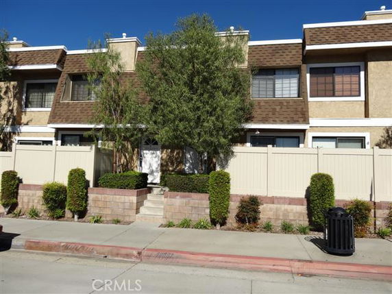 Front view of a two-story townhouse with a brown roof and beige exterior, surrounded by a low fence and shrubbery.