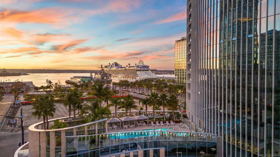 View of modern buildings and a harbor at sunset, featuring palm trees and a cruise ship in the background.