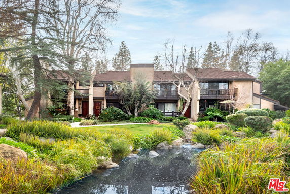 Front view of a two-story building with a brown roof and balconies surrounded by trees and a garden.