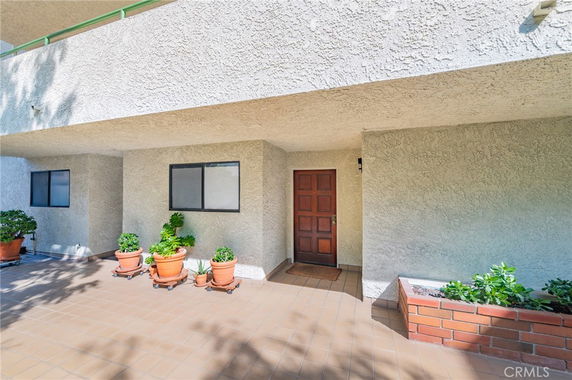 Front view of a building entrance with a wooden door and small windows, featuring potted plants on a tiled patio.