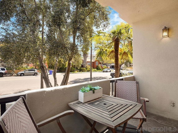 View from a patio with chairs and a small table, overlooking a street with trees and parked cars.