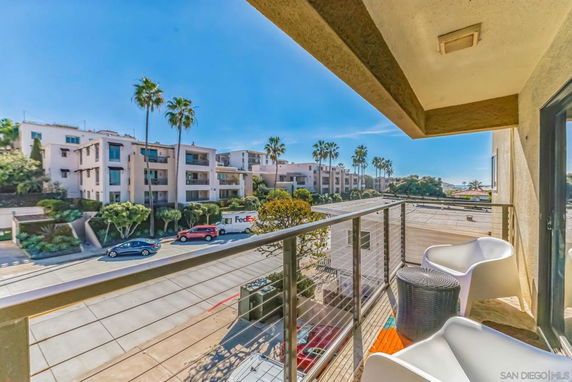 View from a balcony overlooking a street with apartment buildings and palm trees under a clear blue sky.