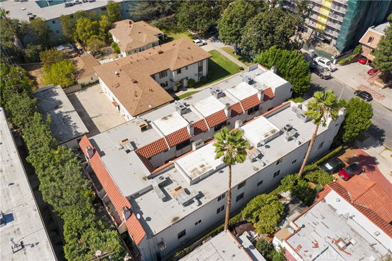Aerial view of residential buildings with flat and gable roofs, surrounded by trees and parked cars.