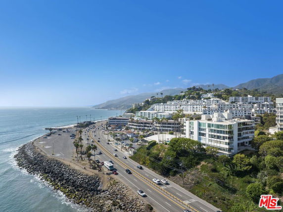 Panoramic view of buildings along a coastal road with ocean and mountain backdrop.
