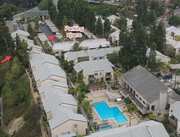 Aerial view of a residential complex with multiple buildings, a swimming pool, and a designated parking area surrounded by trees and greenery.