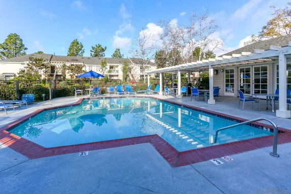 Swimming pool area with seating and a covered patio near apartment buildings.