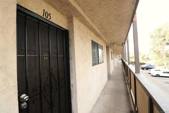 View along an outdoor corridor of a residential building with numbered doors and a parking area visible.