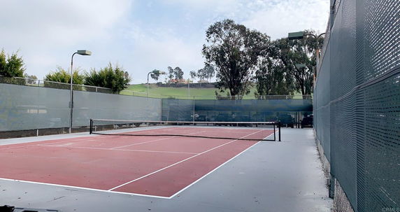 Outdoor tennis court with surrounding fences and trees.