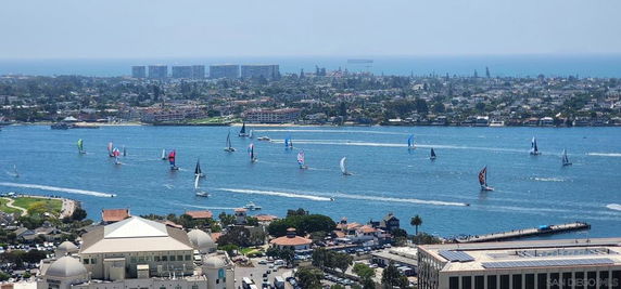 Panoramic view of a coastal area with several sailboats on the water and buildings in the background.