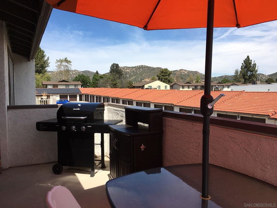 View from a balcony with a grill and outdoor furniture, overlooking rooftops and mountains in the distance.