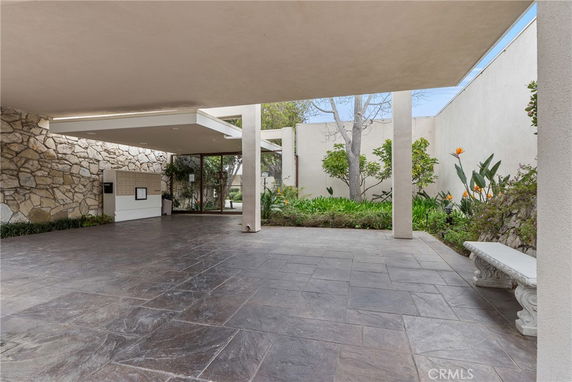 Covered outdoor area with stone walls and tiled flooring, featuring a seating bench and surrounding greenery.