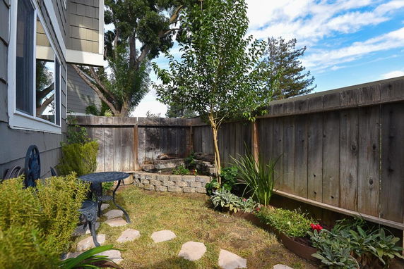 Outdoor garden area with a small patio table, chairs, and plants, adjacent to a building with visible windows.