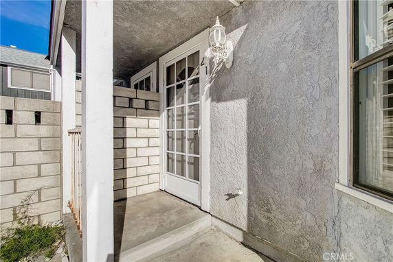 Partial front view of a house with white stucco walls and a glass door entrance. There is a brick wall and a light fixture beside the door.