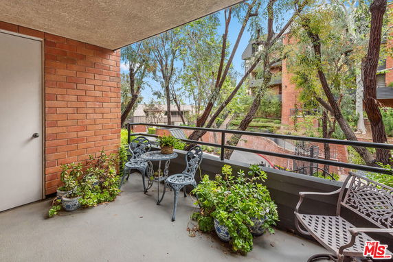 Balcony view with outdoor seating and plants overlooking a courtyard with trees and adjacent buildings.